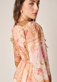 Junge Frau trägt ein rosa-beiges Kleid mit floralem Muster und Rüschen und blickt über ihre Schulter vor einem schlichten Hintergrund.