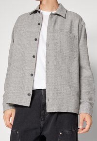 Camicia grigia a maniche lunghe con texture, colletto, taschino frontale e bottoni neri, indossata sopra una maglietta bianca e pantaloni scuri.