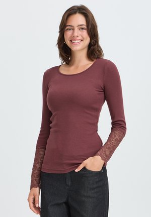 Jeune femme souriante portant un haut moulant bordeaux à manches longues avec des poignets en dentelle et un jean noir, debout avec une main dans la poche.