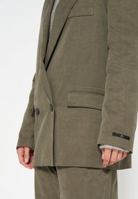 Blazer verde oliva doppiopetto con due bottoni, bavero a punta, tasche laterali e un'etichetta in tessuto nero sul polsino. Materiale testurizzato.