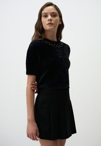 Czarna bluzka z krótkim rękawem z sztucznego futra, z okrągłym dekoltem ozdobionym srebrnymi ćwiekami, w zestawie z czarną plisowaną spódnicą. Prosty i teksturowany design.