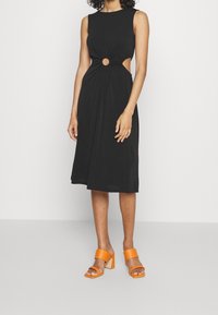 Robe noire sans manches avec un détail découpé comprenant une ouverture ronde, tissu fluide, longueur genou, assortie avec des sandales à talons blocs orange.