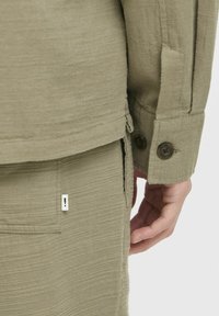 Veste vert olive en tissu texturé, avec des coutures visibles et deux boutons foncés au niveau des poignets ; la main repose sur un pantalon assorti.