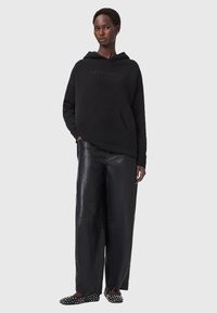 Felpa nera con cappuccio e tasca frontale, abbinata a pantaloni larghi in pelle nera e scarpe nere con pois bianchi.