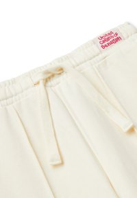 Krémové tepláky zblízka ukazující elastický pás, zavázanou šňůrku, boční kapsu a růžovou nálepku United Colors of Benetton.