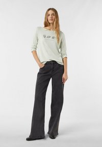 Jasnozielona koszula z długim rękawem z nadrukowanymi ikonami jedzenia i napisem "BREAKFASTCLUB", w połączeniu z czarnymi szerokimi spodniami i czarnymi butami, w pozycji stojącej.