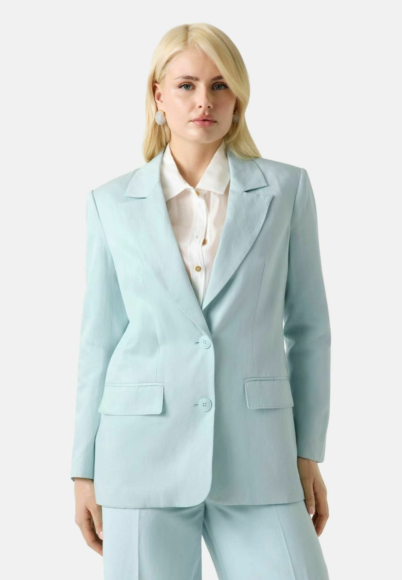 Femme blonde portant un blazer bleu clair sur mesure et un pantalon assorti avec une chemise blanche à boutons sur un fond uni.
