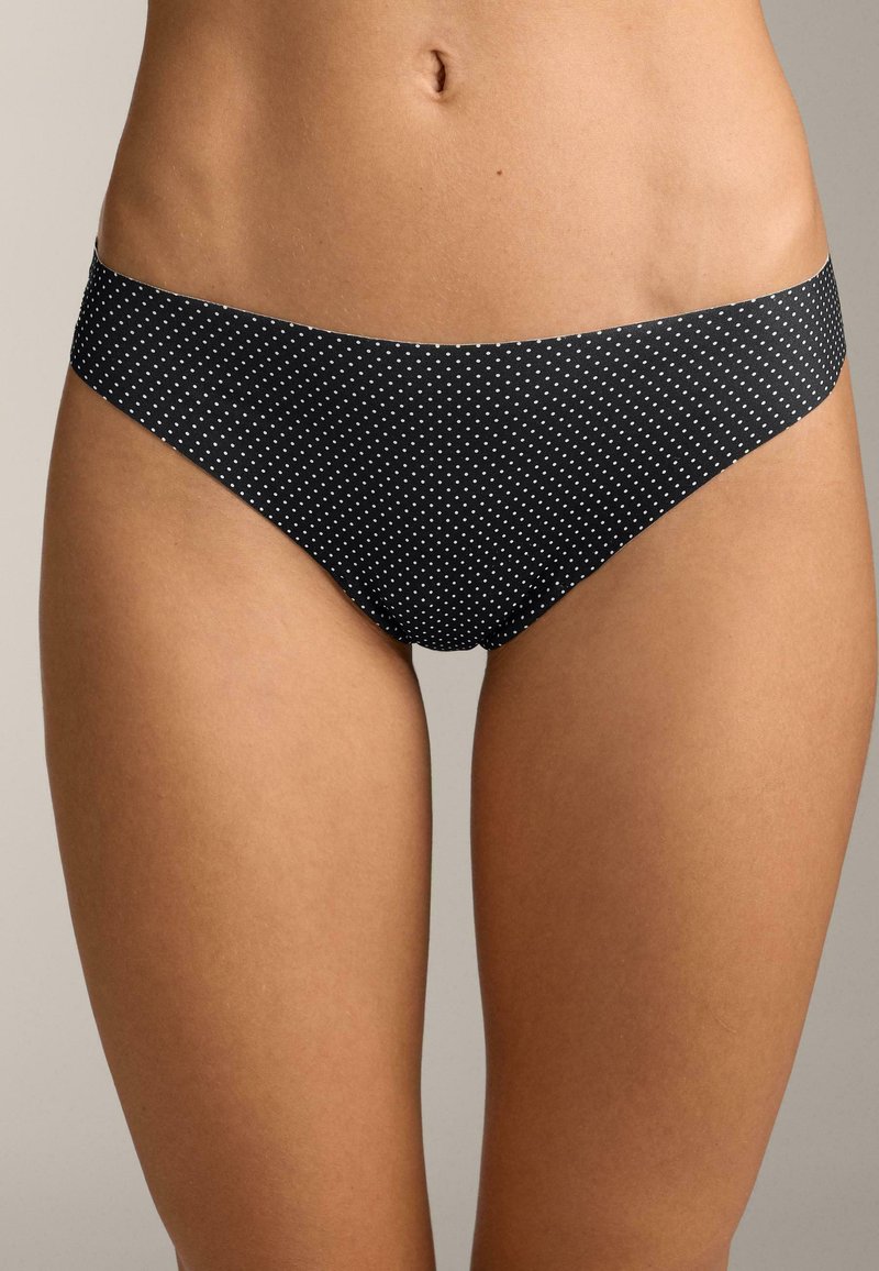 Čierne bikiny nohavičky s malými bielymi bodkami, ktoré má na sebe osoba, zobrazujúce pás, boky a hornú časť stehien na neutrálnom pozadí.