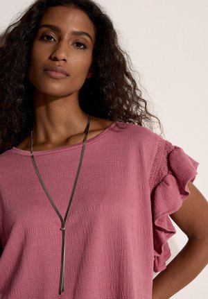 Vrouw met krullend haar, gekleed in een getextureerde roze top met geplooide mouwen en een lange zilveren Y-vormige ketting tegen een lichte achtergrond.