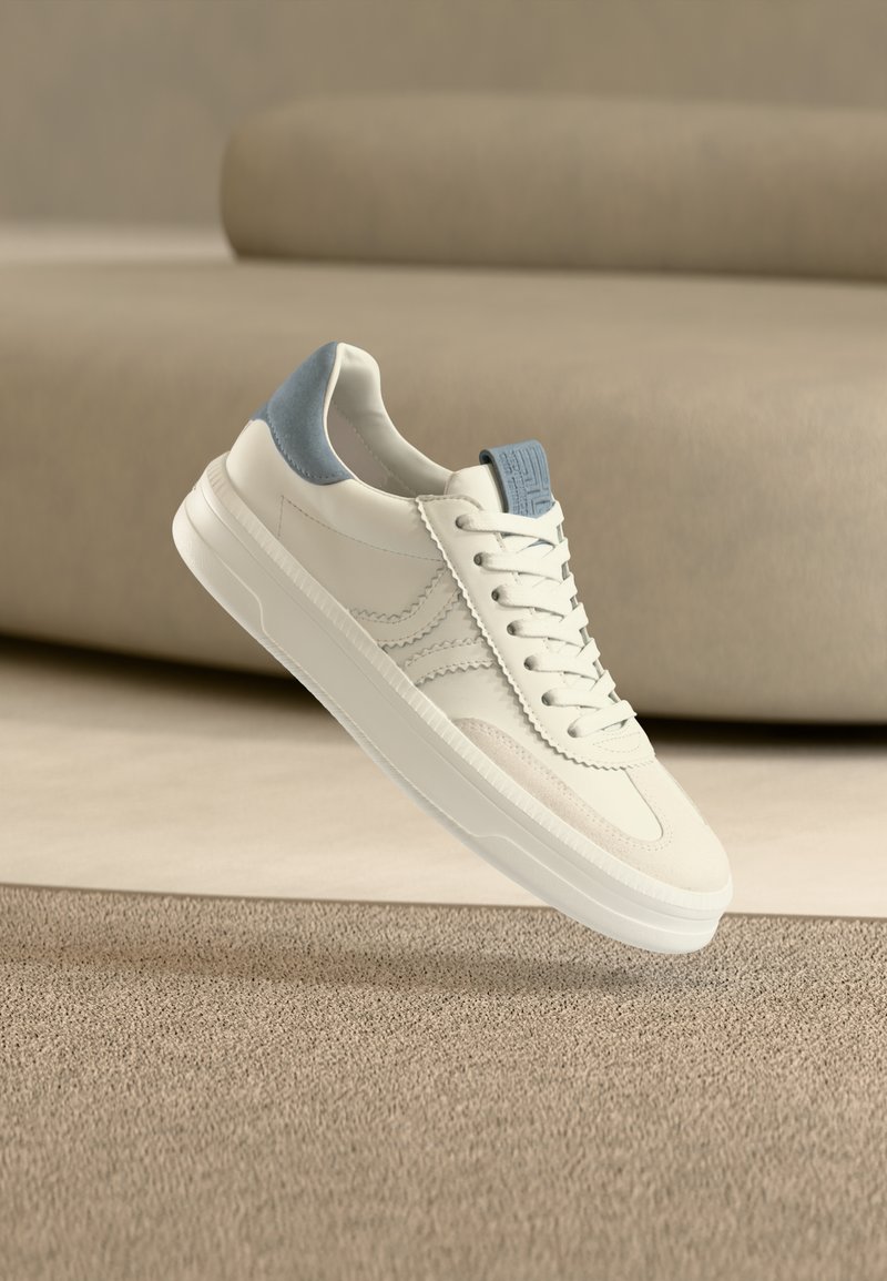 Λευκό αθλητικό παπούτσι με λεπτομέρεια στο τακούνι σε ανοιχτό μπλε και κορδόνια στο μπροστινό μέρος, να αιωρείται πάνω από μπεζ χαλί με θολό φόντο ένα καναπέ μπεζ χρώματος.