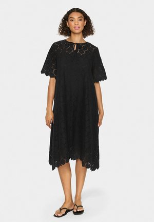 Black lace dress with short flared sleeves, scalloped hem, keyhole neckline, and floral pattern details. Worn with black flip-flops.