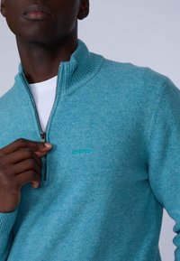 Pull léger bleu, zippé, avec un col haut et une texture côtelée. Il présente un petit logo brodé sur la poitrine et une couche intérieure blanche.