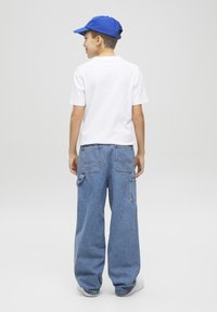 Maglietta bianca a maniche corte, cappellino blu, pantaloni oversize in denim blu con grandi tasche e scarpe da ginnastica bianche, visti da dietro.