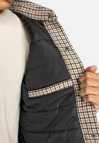 Osoba trzyma otwartą brązowo-białą kurtkę w wzór houndstooth, pokazując jej czarne pikowane podszycie i kieszeń.