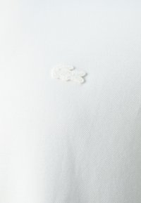Tessuto bianco testurizzato con un logo ricamato a forma di coccodrillo. Il design è minimale e pulito, incentrato sulla semplicità.