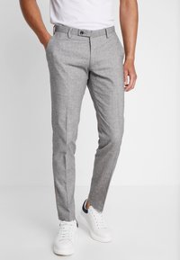 Muž v úzkých šedých kalhotách a bílých teniskách, stojící s jednou rukou v kapse na čistě bílém pozadí.