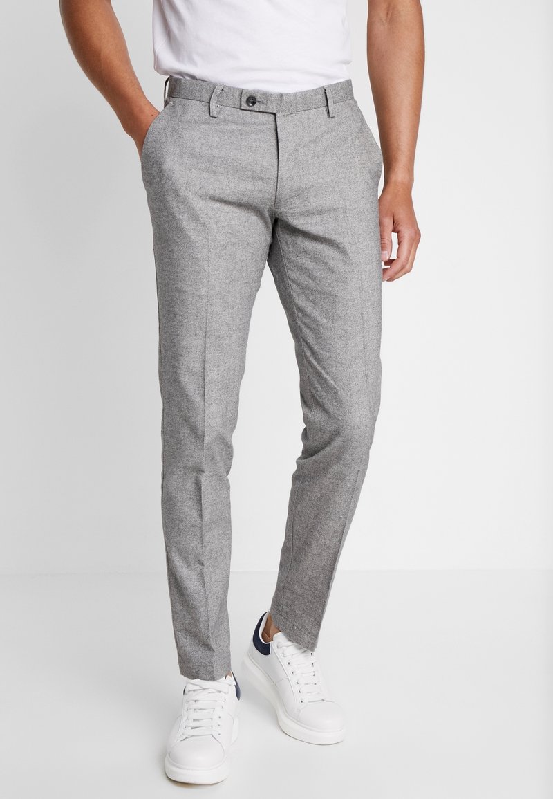 Muž v úzkých šedých kalhotách a bílých teniskách, stojící s jednou rukou v kapse na čistě bílém pozadí.