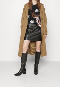 Cappotto trench marrone sopra a un maglione nero con renna, abbinato a una gonna in pelle nera e stivali alti neri con tacchi quadrati.