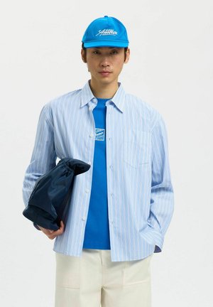 Mann mit blauem Cap, blauem Grafik-T-Shirt, hellblau gestreiftem Hemd mit Knöpfen, beigen Hosen, hält eine dunkelblaue Jacke in einer Hand.