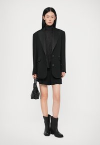 Zwarte blazer met satijnen revers, gecombineerd met een donkere coltrui en shorts. Accessoires zijn een kleine zwarte tas met een netpatroon en zwarte enkellaarsjes.