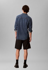 Mann steht mit dem Rücken zur Kamera, trägt ein hochgekrempeltes Jeanshemd, schwarze Shorts, schwarze Socken und schwarze Business-Schuhe vor schlichtem grauem Hintergrund.