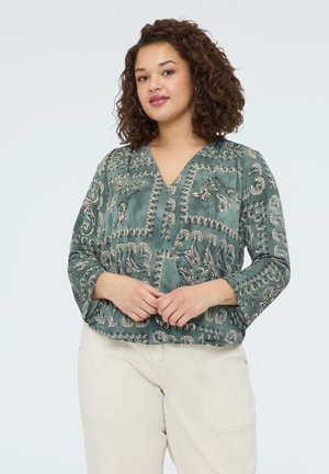 Kiharatukkainen nainen, joka pukeutunut vihreään paisley-kuvioiseen puseroon ja vaaleanbeigeihin housuihin, seisoo yksinkertaisen vaalean taustan edessä.