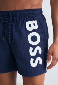 Jūroje mėlynos maudymosi šortai su elastiniu juosmeniu ir raišteliu. Šone yra dideli balti "BOSS" užrašai. Lengvas, tekstūruotas audinys.