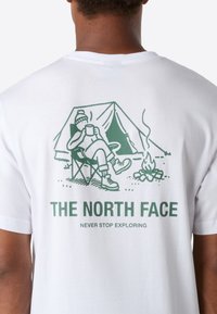 Osoba v bílém tričku s zelenou grafikou kemperu, který pije u táboráku před stanem, s textem "THE NORTH FACE" a "NEVER STOP EXPLORING."