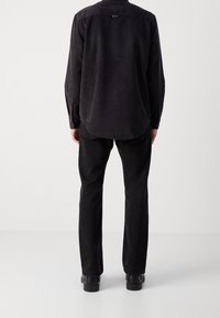 Camisa negra de manga larga hecha de tela texturizada, combinada con pantalones negros de pierna recta y zapatos negros. La vista desde la parte trasera muestra la longitud y el ajuste de la camisa.