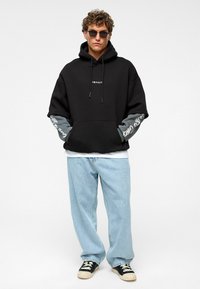 Zwarte oversized hoodie met grijze mouwen, witte 'IMPACT' logo en voorzak; gecombineerd met lichtblauwe losse jeans en zwarte sneakers.