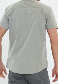 Camisa de manga corta en tejido gris claro, con un cuello clásico, yugo en la parte trasera y dobladillo curvado. Textura suave, sin patrones visibles.