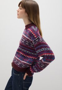 Strickpullover mit einem lila, roten und blauen Fair Isle-Muster, rundem Halsausschnitt und gerippten Bündchen, kombiniert mit dunklen Jeans.