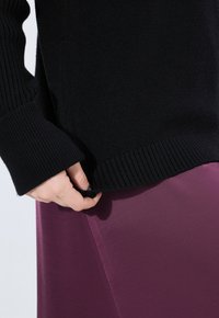 Mano che aggiusta l'orlo di un maglione nero a coste indossato sopra una gonna o un vestito viola, mostrando la texture del tessuto e la vestibilità.