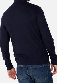 Mörkblå, stickad tröja med hög krage och ribbmuddar. Har en ren baksidesdesign och bärs över blå jeans.