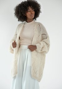 Beżowy, dziany kardigan z otwartym przodem i luźnymi rękawami, z wzorem w diamenty i miękkim, teksturowanym wykończeniem. W parze z lekką bluzką i szerokimi spodniami.