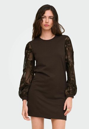 Femme portant une robe noire à manches longues avec des manches transparentes brodées de fleurs, debout devant un fond clair uni.