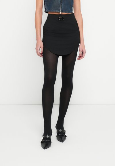 Mini-jupe noire à ourlet incurvé, portée sur des collants noirs transparents, et des talons pointus noirs ornés de détails en boucle argentée. Haut court en denim.