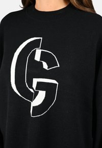 Pull en maille noir avec un grand design graphique blanc représentant des lettres 'C' et 'G' qui se chevauchent. Encolure ronde et poignets côtelés. Texture douce.