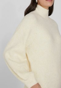 Suéter de color crema con cuello alto simulado, textura acanalada, mangas gruesas y un ajuste relajado, hecho de material de punto suave.