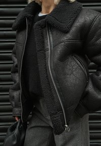 Veste en shearling noir avec un large col, surface texturée, fermeture éclair à l'avant et grandes poches, associée à un pull en laine et un pantalon en chevrons.