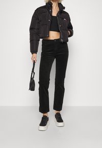 Zwarte pufferjack met een cropped ontwerp, gecombineerd met zwarte corduroy broeken. De sneakers hebben een witte zool en de tas is klein en zwart.