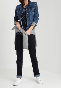 Epämuodollinen asu, johon kuuluu farkkutakki, tummansininen hihaton paita, jossa on valkoisia kuvioita, harmaa neule vyötäröllä, mustat farkut ja valkoiset lenkkarit. Rento istuvuus.