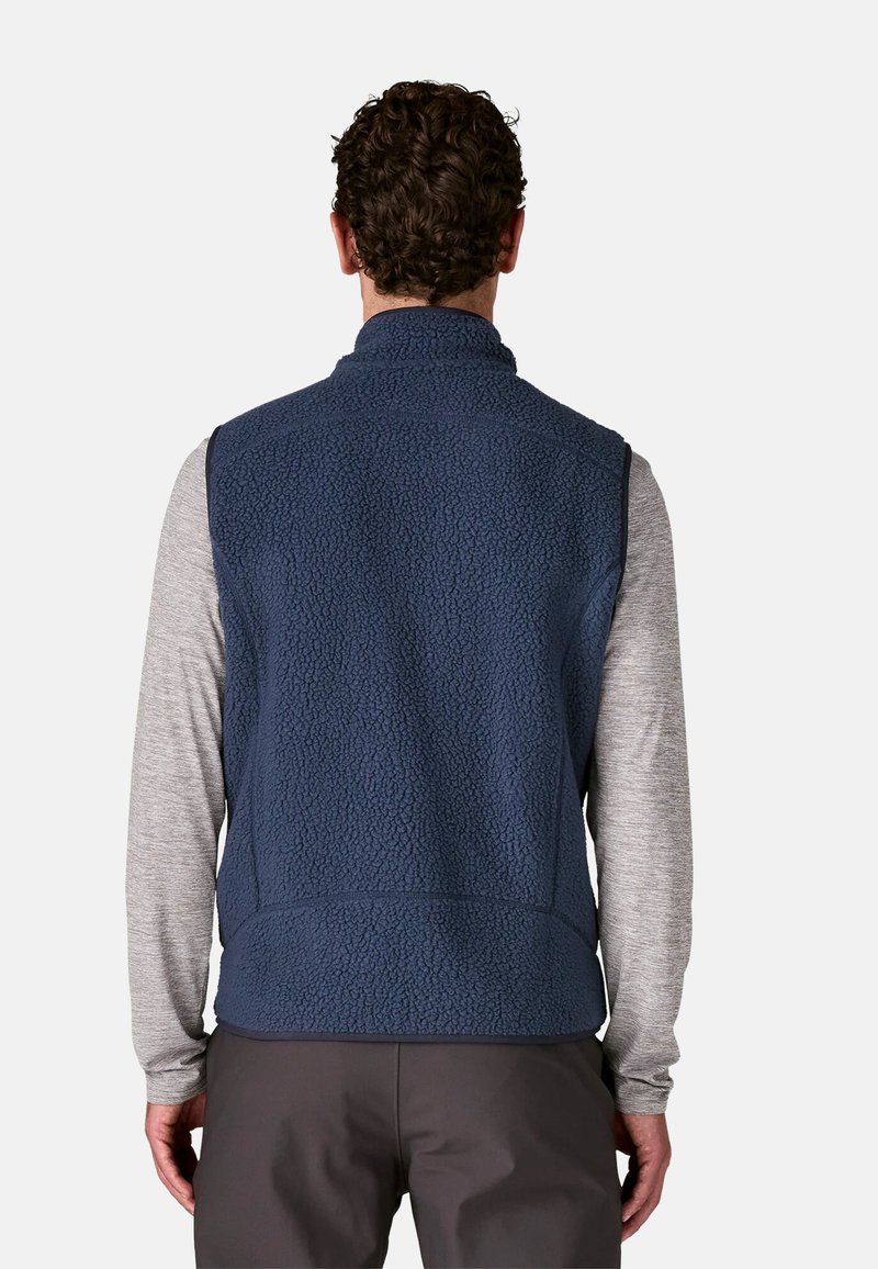 Gilet in pile blu navy con finitura testurizzata, colletto alto e senza maniche. Indossato sopra una camicia a maniche lunghe grigia chiara.