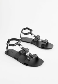 Černé kožené sandály s plochou podrážkou, které mají více pásků, kulaté kovové detaily a nastavitelné uzávěry kolem kotníků.