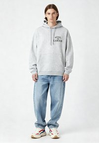 Szürke kapucnis pulóver, oversize szabású, zsinórral ellátott kapucnival és elülső zsebbel. Fekete nyomtatott szöveg: "HÔTEL DE L'AMOUR." Világoskék farmernadrággal és sportcipővel viselve.