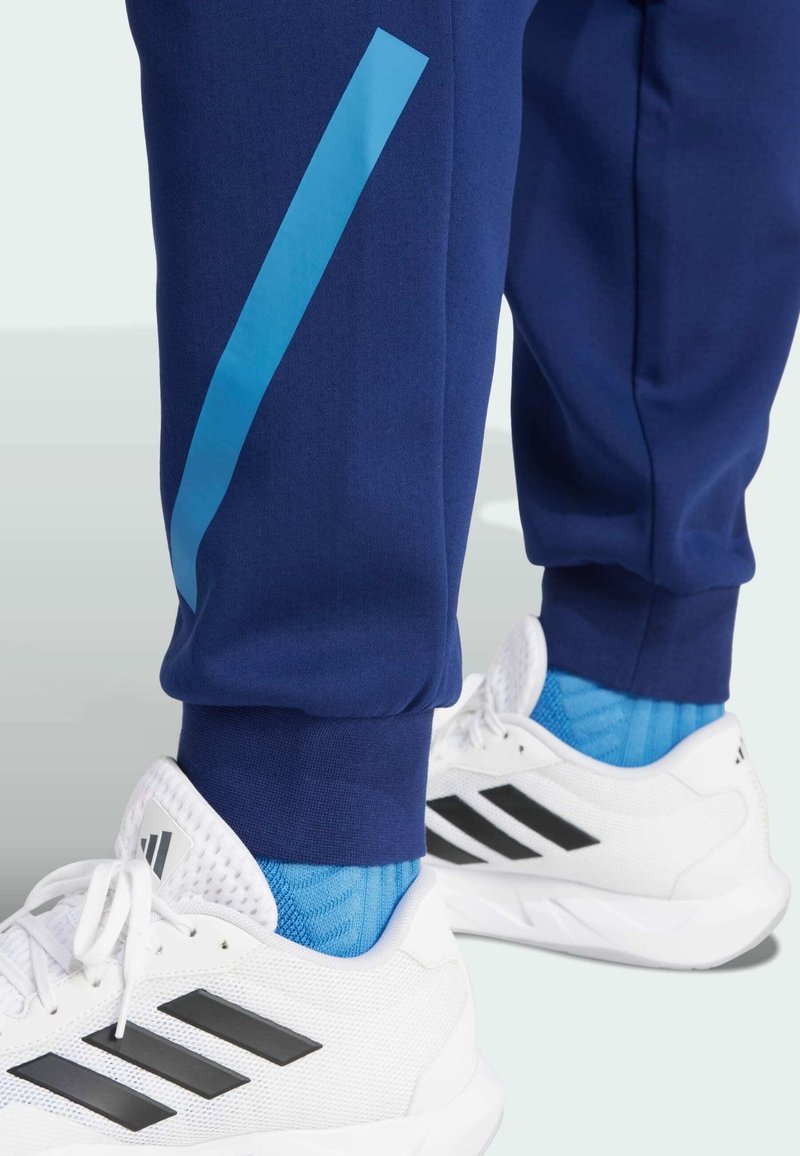Marineblaue Jogginghose mit einem blauen vertikalen Streifen. Der Stoff ist glatt und hat gerippte Bündchen. Kombiniert mit weißen Sportschuhen mit schwarzen Akzenten.