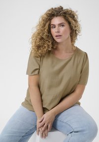 Blouse à manches courtes, vert olive, avec une coupe décontractée et un tissu texturé, associée à un jean bleu clair. Encolure douce et détails subtils aux épaules.