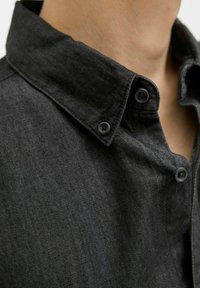 Cămașă din denim negru cu guler cu nasturi, două nasturi negri și un material texturat. Viziune de aproape care evidențiază detaliile cămășii.