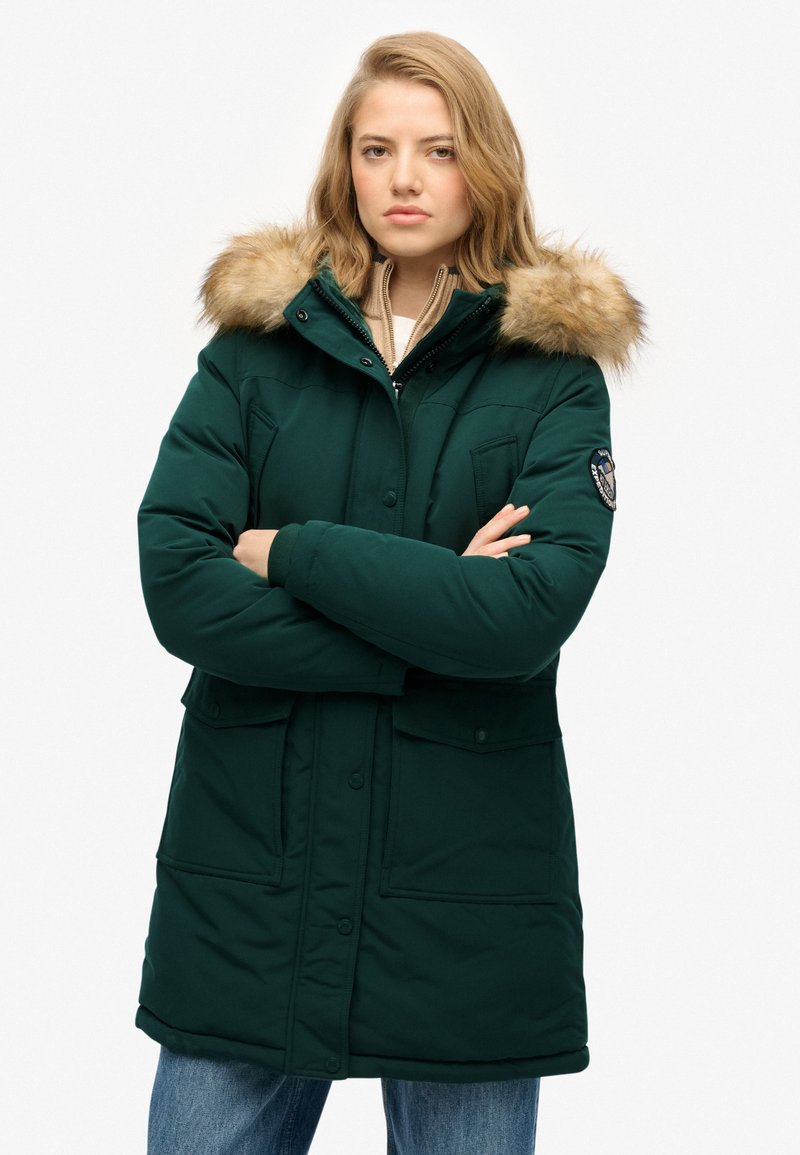 Parka de color verde oscuro con borde de pelo sintético en la capucha, botones a presión, dos bolsillos frontales y un ajuste relajado, combinada con unos jeans de color azul claro.