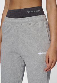 Grå joggingbukser med en mørkegrå elastisk linning. Har en hvid logo på venstre side og et blødt, tekstureret stof.